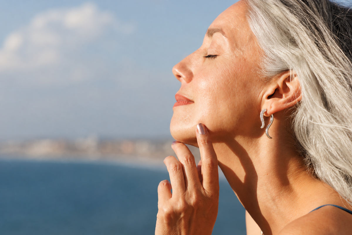 Model wearing sculptural Premium Pavé Swoop earrings with sparkling diamonds and curved silver forms, against a softly blurred ocean backdrop.